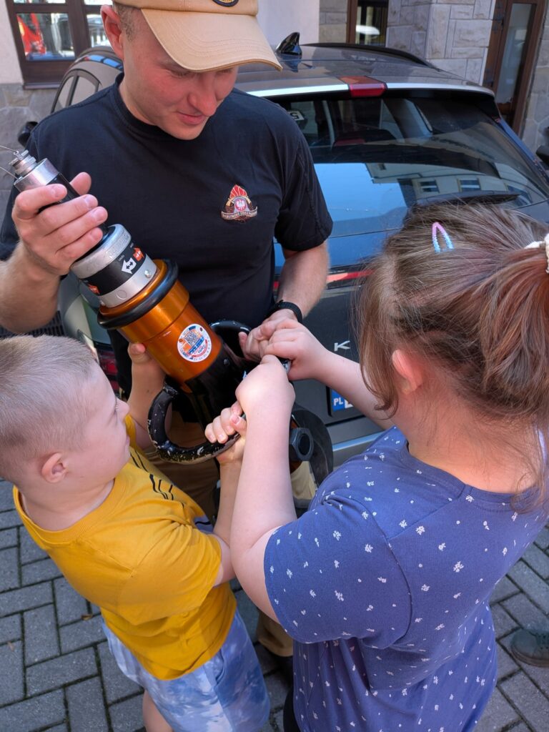 Strażak w czarnej koszulce i beżowej czapce pomaga dwójce dzieci wspólnie utrzymać ciężkie, hydrauliczne narzędzie ratownicze.