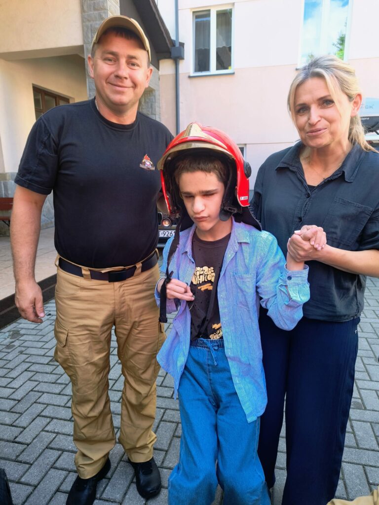 Chłopiec w dużym, czerwonym hełmie strażackim stoi na brukowanym podwórku pomiędzy uśmiechniętym strażakiem a kobietą trzymającą go za rękę.
