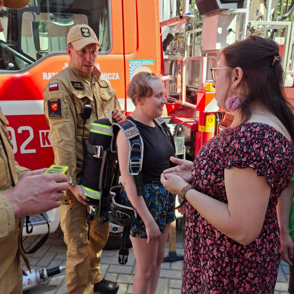 Dziewczyna przymierza profesjonalny aparat oddechowy butlowy, podczas gdy asekuruje ją strażak, a obok rozmawia z nią kobieta w kwiecistej sukience.