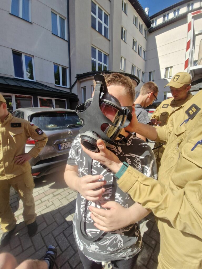 Strażak w beżowym mundurze pomaga chłopakowi dopasować i założyć na twarz czarną maskę przeciwgazową.