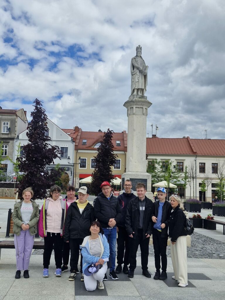  Grupa osób pozuje do wspólnego zdjęcia na miejskim placu przed wysokim pomnikiem przedstawiającym postać króla na kolumnie. W tle widoczne są kamienice z czerwonymi dachami.