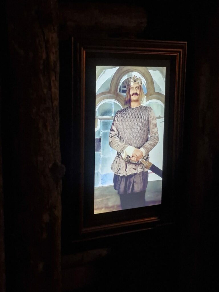Pionowy ekran w ciemnym pomieszczeniu, na którym wyświetlany jest cyfrowy portret wąsatego mężczyzny w stroju stylizowanym na średniowiecznego rycerza lub króla z koroną i mieczem.