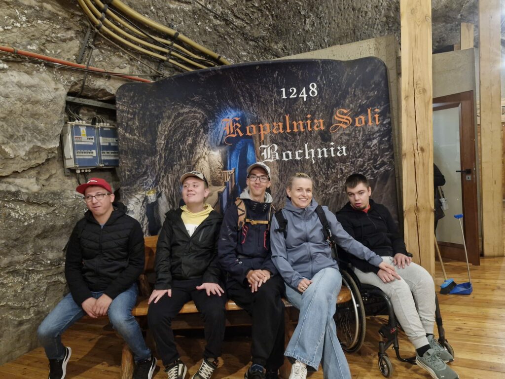  Pięć osób siedzi na drewnianej ławce i pozuje do zdjęcia na tle pamiątkowej ścianki z napisem „1248 Kopalnia Soli Bochnia”. Po prawej stronie siedzi chłopak na wózku inwalidzkim.