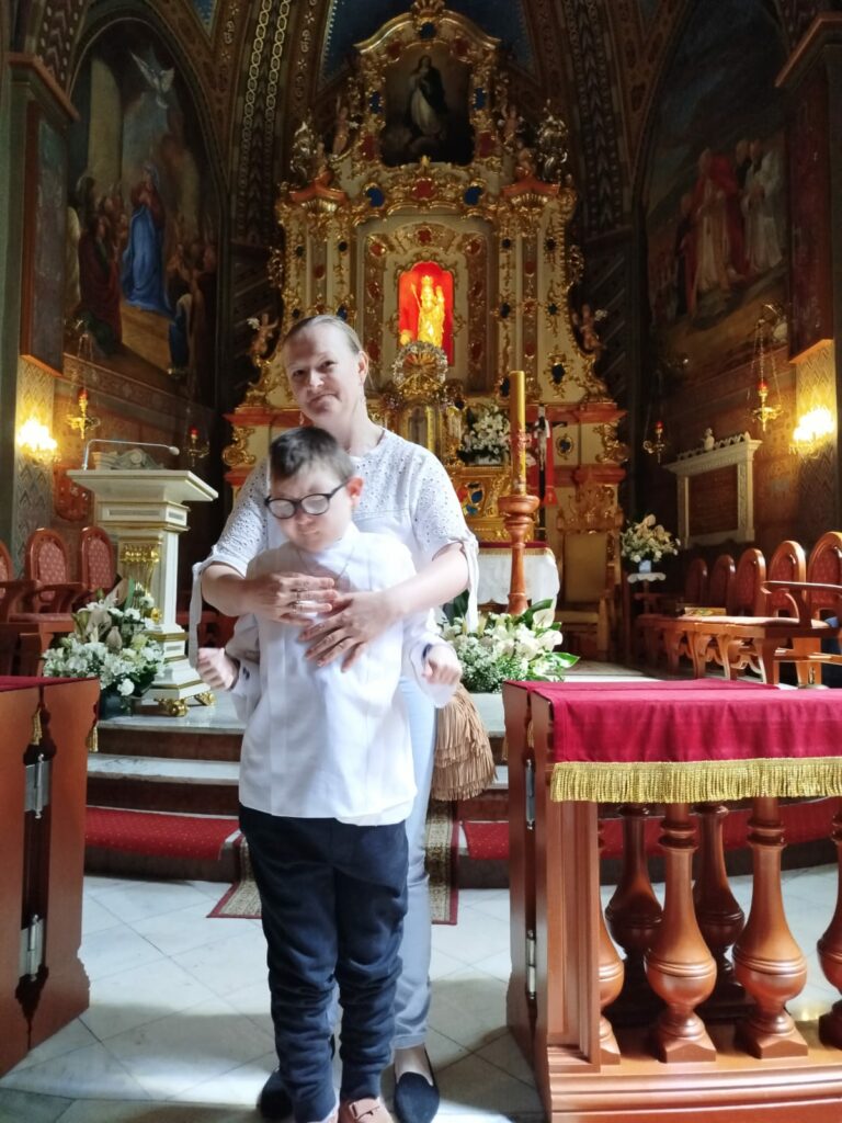 Kobieta i chłopiec w okularach stoją we wnętrzu bogato zdobionego kościoła przed złotym ołtarzem. Kobieta obejmuje chłopca od tyłu.
