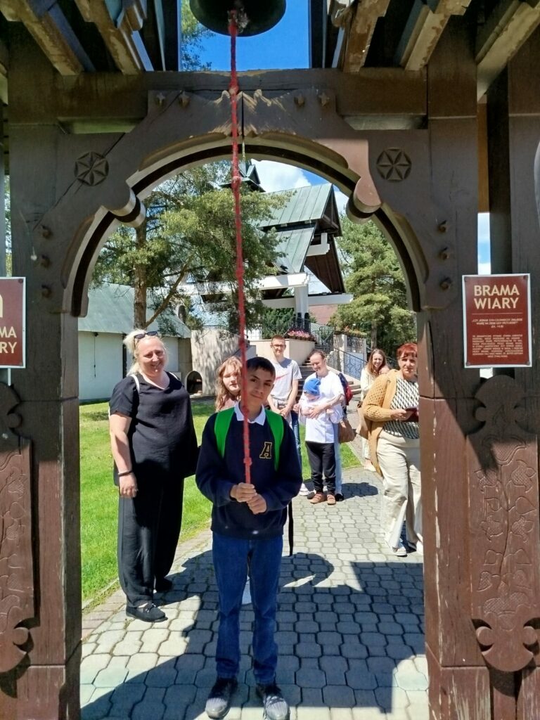 Chłopiec w ciemnej bluzie z zielonym plecakiem stoi pod drewnianą bramą z napisem „Brama Wiary” i trzyma czerwony sznur od dzwonu. W tle stoją inni ludzie.