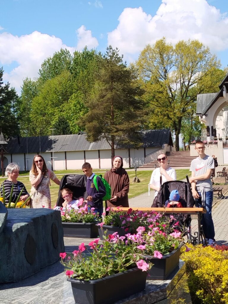 Grupa osób, w tym dzieci i dorosłe kobiety, pozuje do zdjęcia w ogrodzie przed budynkiem. Na pierwszym planie widać podłużne donice z różowymi kwiatami.
