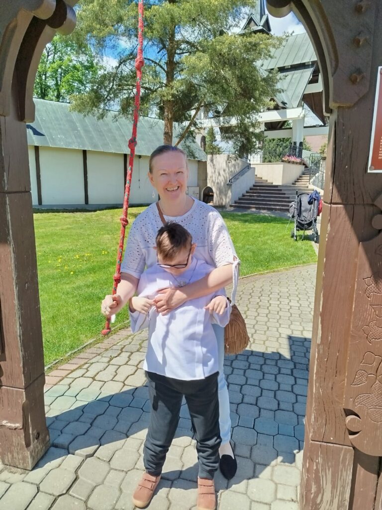 Uśmiechnięta kobieta stojąca pod drewnianą bramą obejmuje od tyłu chłopca, pomagając mu trzymać czerwony sznur zwisający z góry.