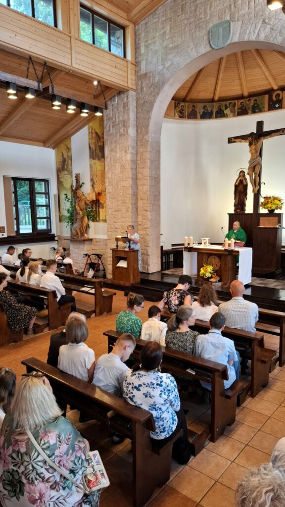 Wnętrze kaplicy o jasnych, kamienno-drewnianych ścianach. W ławkach siedzą wierni przodem do ołtarza, przy którym stoi ksiądz w zielonym ornacie. Kobieta stoi przy ambonie po lewej stronie i czyta tekst. W tle widać duży krucyfiks.