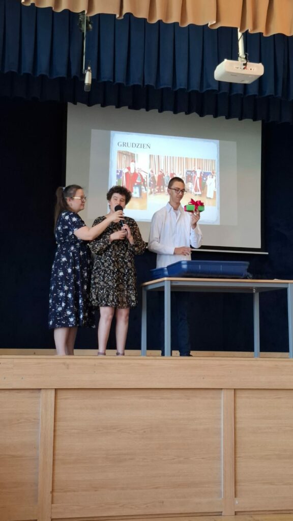 Na scenie stoją trzy osoby pod dużym ekranem z napisem „GRUDZIEŃ”. Kobieta w ciemnej sukience w kwiaty trzyma mikrofon dla śpiewającej dziewczyny. Obok nich stoi chłopak w białej koszuli, trzymający w dłoni małe, zielone pudełko z czerwoną kokardą.