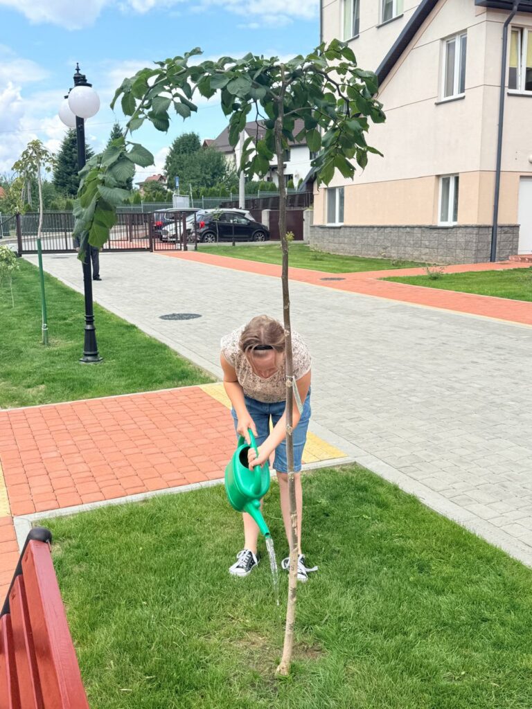Osoba w beżowej koszulce w kwiaty i niebieskich spodenkach pochyla się na trawniku, podlewając z zielonej konewki pień młodego drzewka o parasolowatej koronie. W tle widać chodnik, latarnię, samochody i budynek mieszkalny.