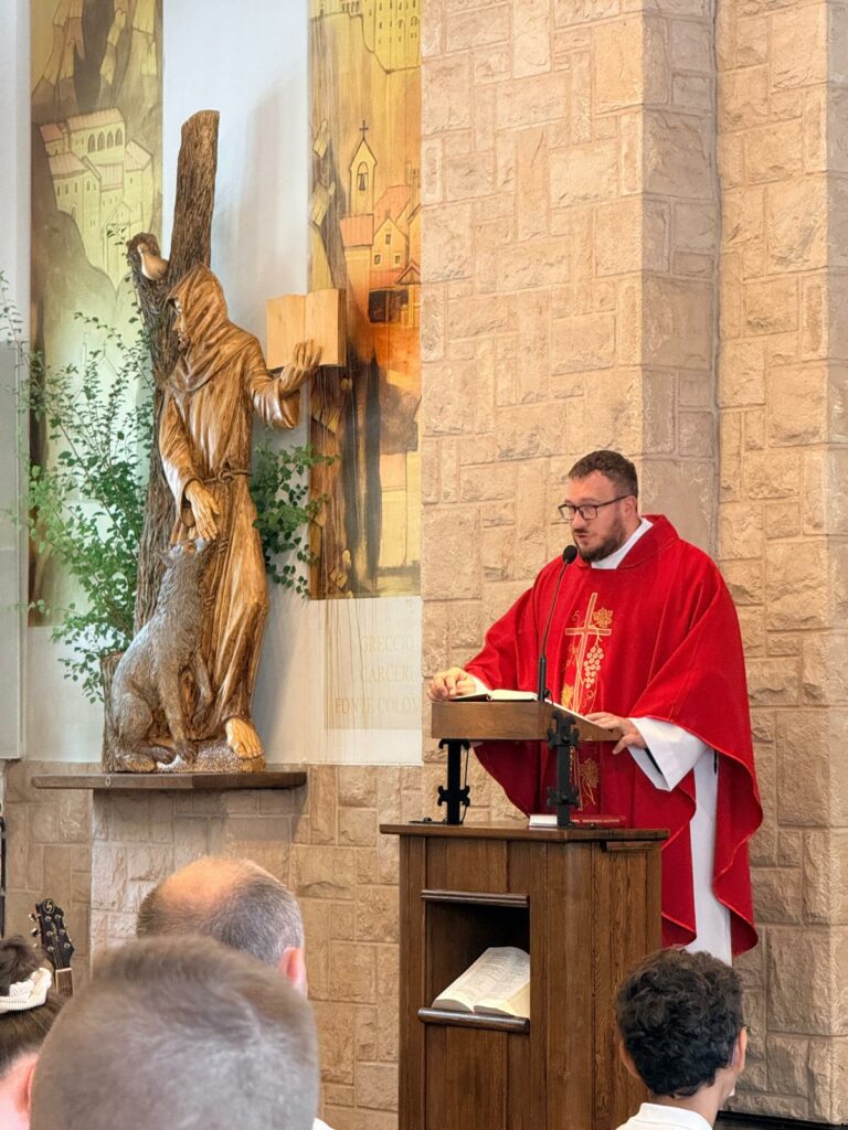 Ksiądz w czerwonych szatach liturgicznych przy ambonie, obok drewniana rzeźba świętego.