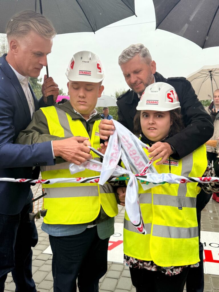 Zbliżenie na młodą kobietę i mężczyznę w kaskach oraz kamizelkach odblaskowych, którzy przy pomocy osób w garniturach rozwiązują wstęgę.