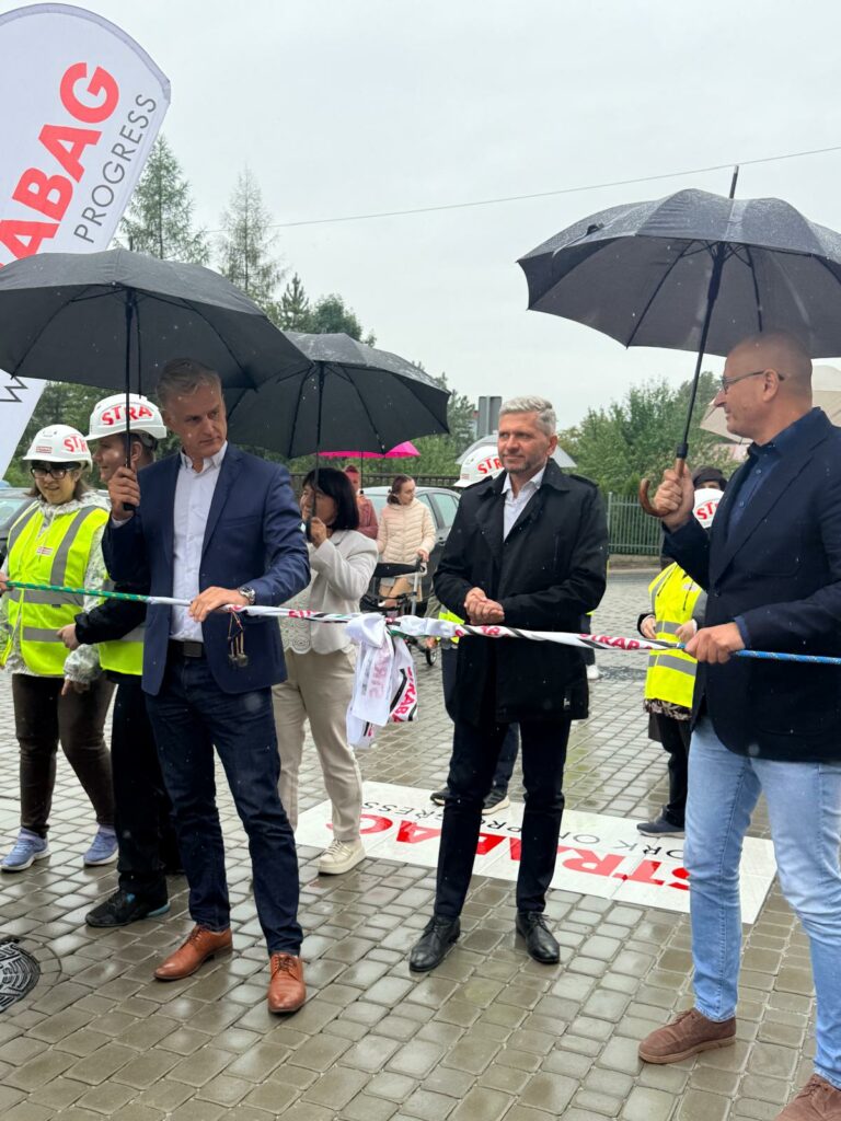 Mężczyźni w garniturach oraz osoby w żółtych kamizelkach odblaskowych i kaskach trzymają wstęgę podczas oficjalnego otwarcia w deszczu pod parasolami.