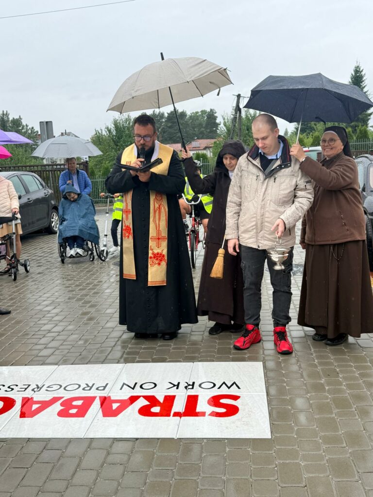Ksiądz ze stułą czyta z księgi podczas uroczystości na zewnątrz w deszczowy dzień, obok niego zakonnice i mężczyzna trzymający kropidło oraz naczynie z wodą święconą.