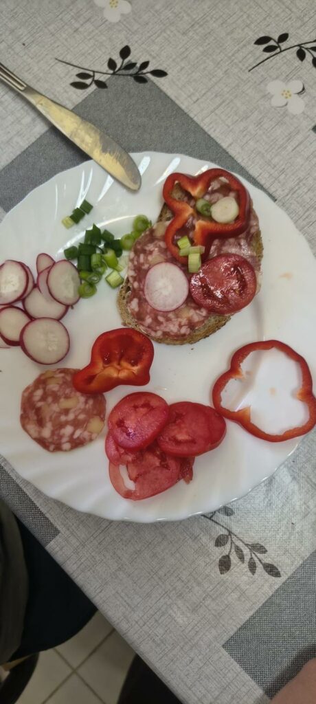 Widok z góry na biały talerz z przygotowaną kanapką z salami, na której ułożono plasterki rzodkiewki, papryki, szczypiorku i pomidora. Obok na talerzu leży pokrojona papryka, pomidory i szczypiorek.