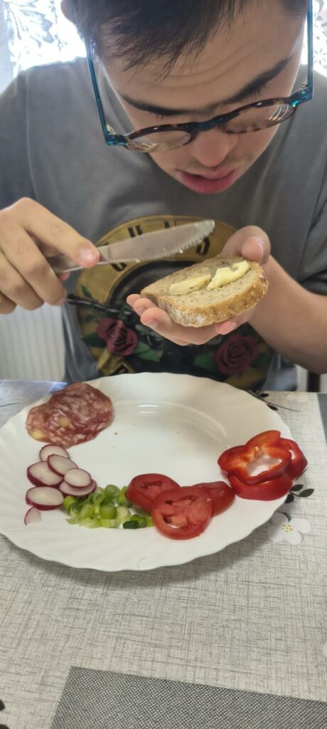 Młody mężczyzna w okularach, rozsmarowuje nożem masło na kromce ciemnego pieczywa. Na białym talerzu przed nim znajdują się pokrojone warzywa (rzodkiewka, szczypiorek, papryka, pomidor) i salami