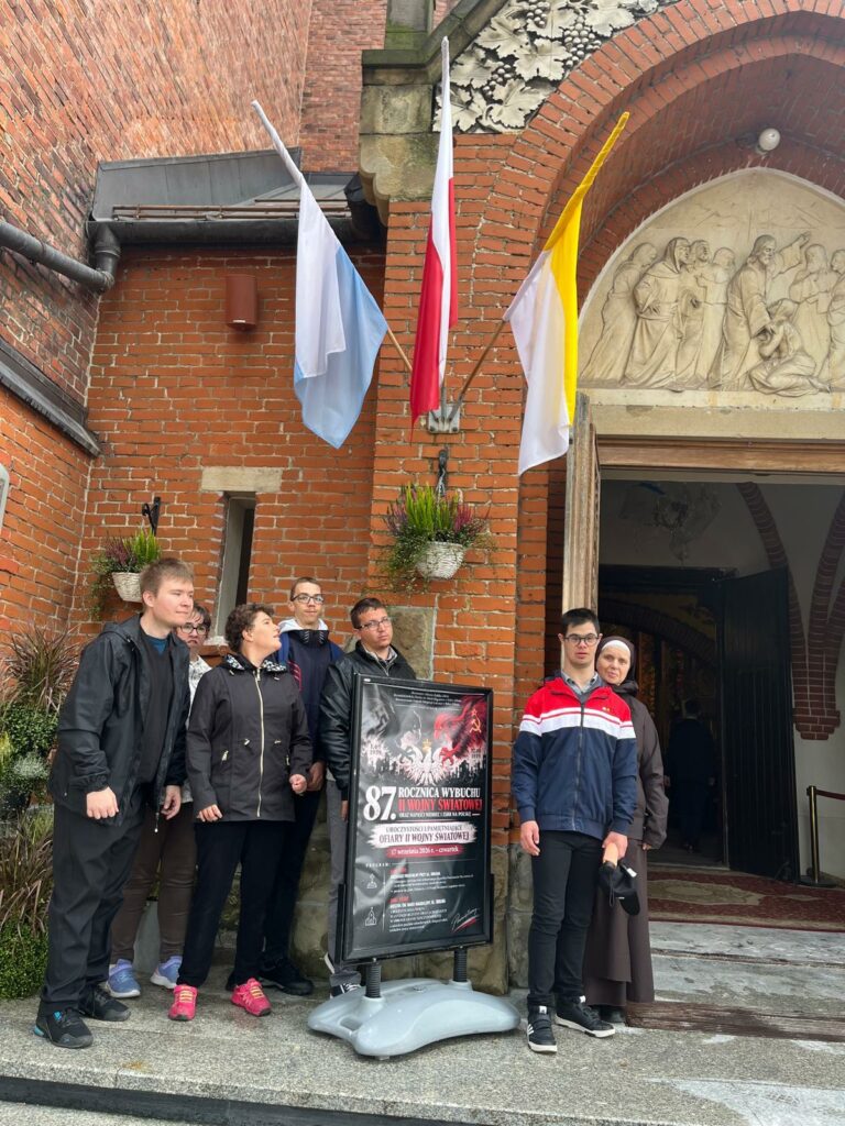 grupa ludzi pozuje do zdjęcia przed wejściem do ceglanego kościoła obok plakatu upamiętniającego rocznicę wybuchu II wojny światowej.
