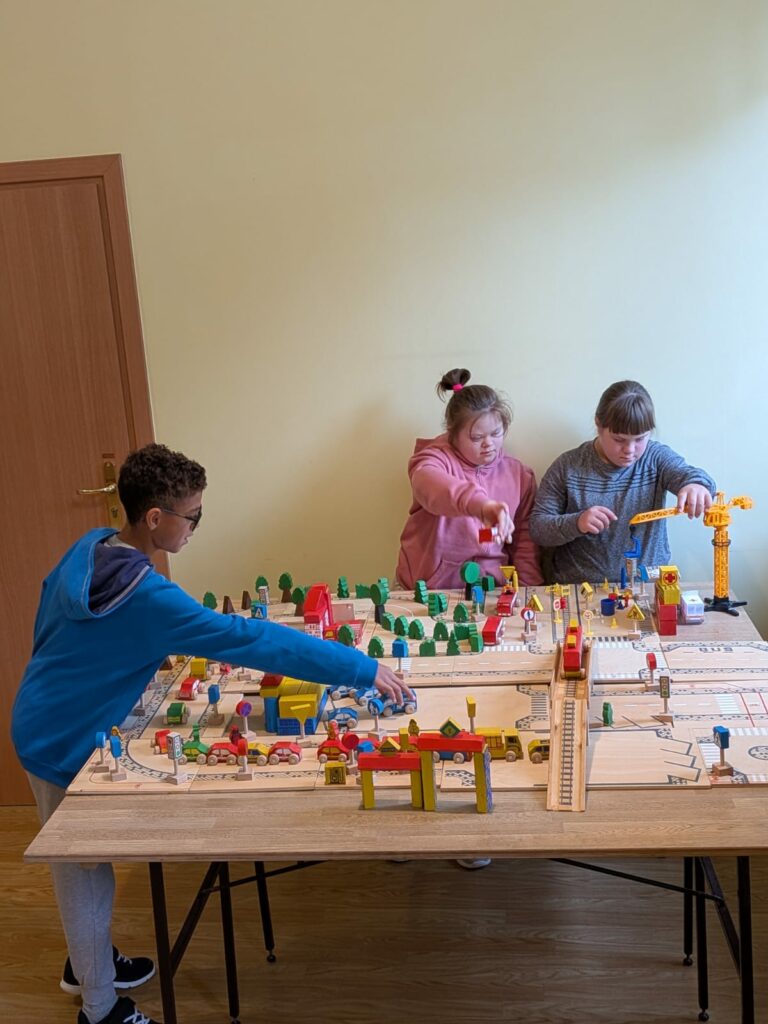 Trójka nastolatków bawi się dużą makietą miasta. Na planszy znajdują się liczne kolorowe klocki, figurki drzew i pojazdów.