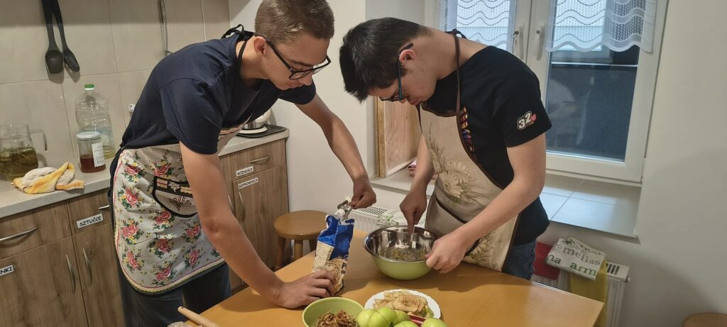 Dwaj młodzi mężczyźni w fartuchach przygotowują w kuchence posiłek. Jeden z nich miesza coś w misce a drugi dosypuje mu składniki.