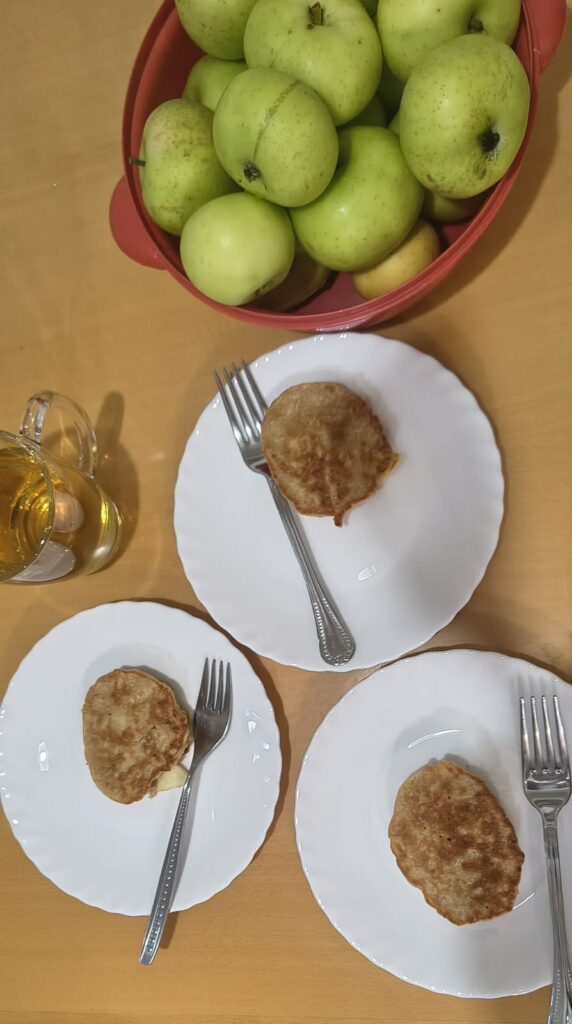 Na stole stoją miska pełna jabłek, szklanka z napojem oraz 3 talerze z plackiem i widelcem.