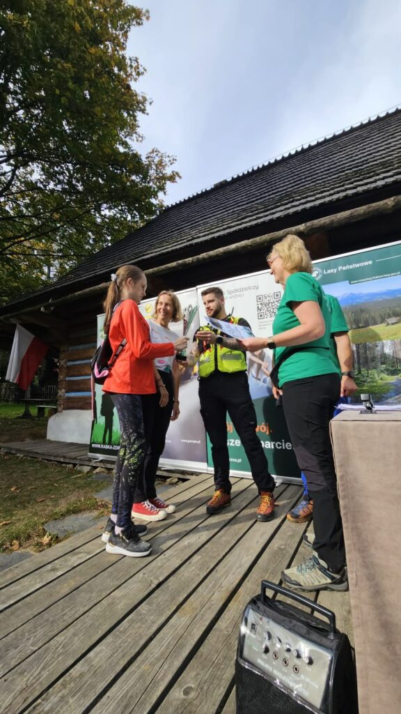 Wręczenie dyplomu i pucharu na drewnianym podeście przed drewnianym budynkiem.