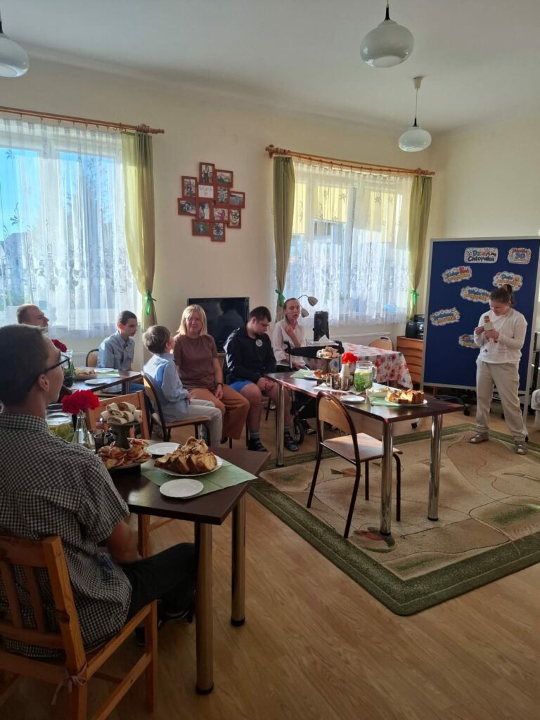 Uczniowie i nauczyciele siedzą przy wspólnym stole i oglądają występ młodej kobiety śpiewającej do mikrofonu..
