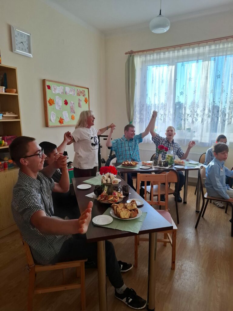 Uczniowie i nauczyciele siedzą przy wspólnym stole podczas spotkania z okazji Dnia Chłopaka, trzymają się za uniesione ręce i śpiewają.