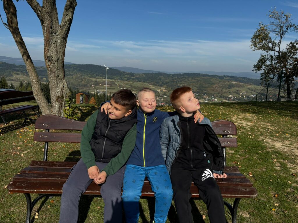 Trzech uczniów siedzi obok siebie na drewnianej ławce, obejmując się ramionami. W tle widać górski krajobraz.
