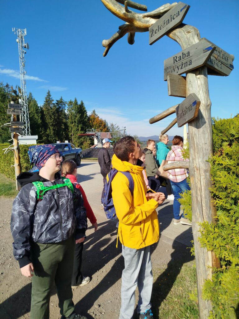 Uczniowie podczas wycieczki oglądają drewniane drogowskazy wskazujące kierunki do pobliskich miejscowości. W tle widać las i górski krajobraz.