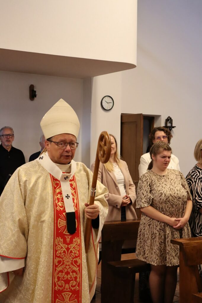 Biskup w szatach liturgicznych z pastorałem stoi w kościele obok zgromadzonych uczestników uroczystości.