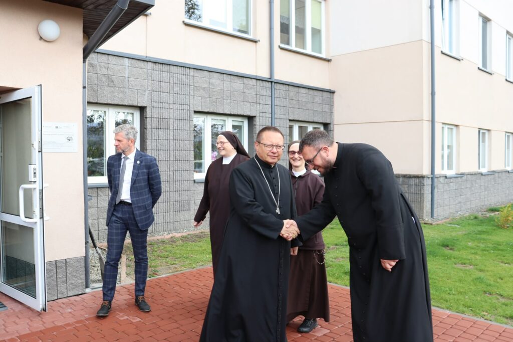 Duchowni witają się przed wejściem do budynku ośrodka, podając sobie ręce w obecności sióstr zakonnych i gości.
