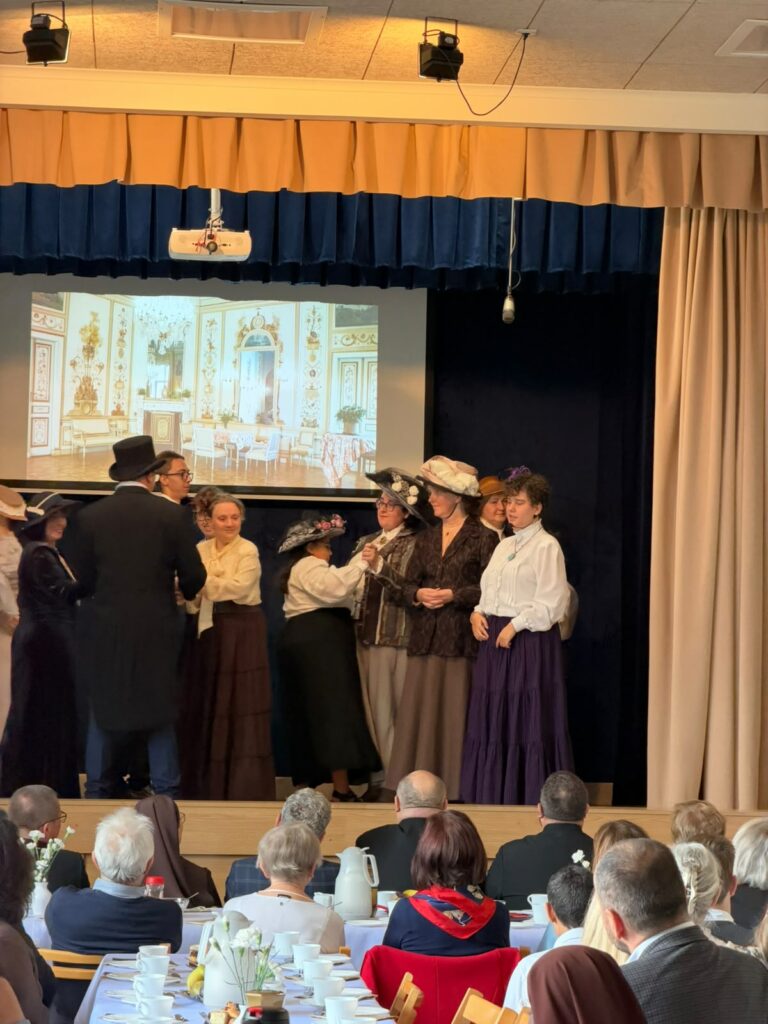 Osoby w strojach historycznych prezentują scenę teatralną na tle dekoracji i wyświetlanej prezentacji.