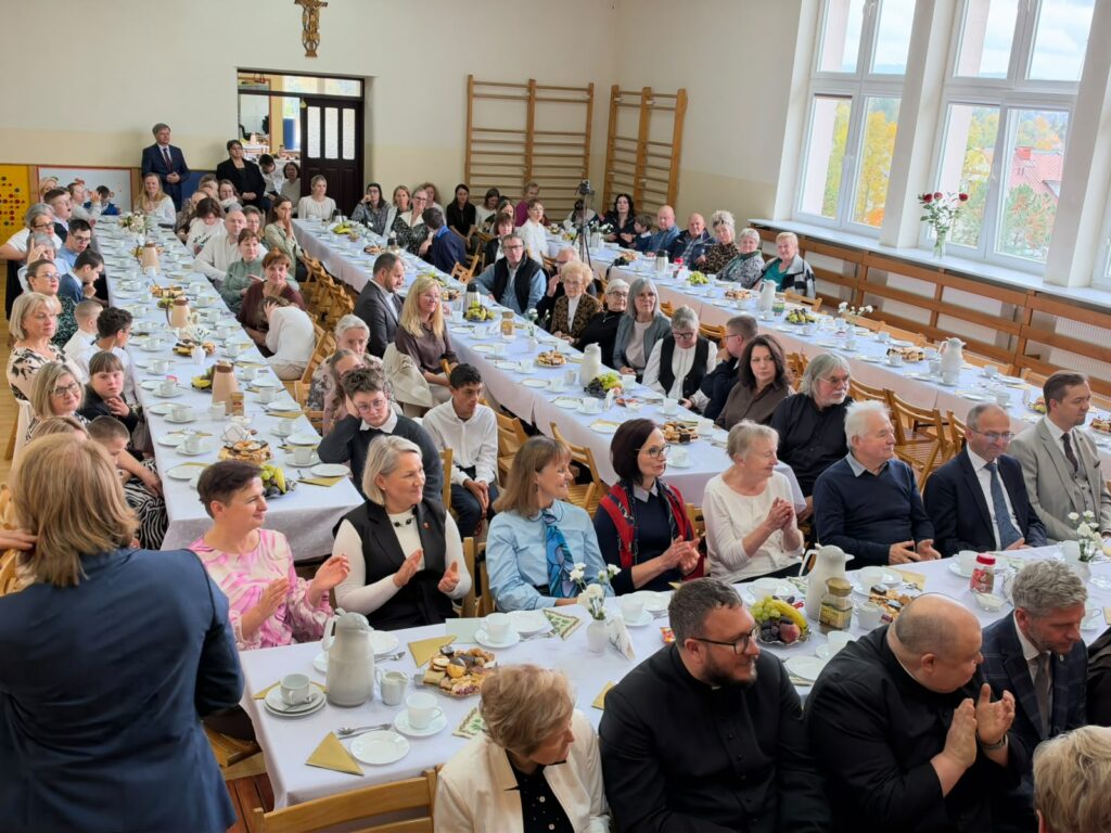 Goście siedzą przy nakrytych stołach w dużej sali, uczestnicząc w uroczystym spotkaniu.