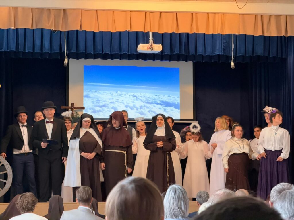 Grupa uczniów i dorosłych w strojach zakonnych, anielskich i historycznych stoi na scenie podczas przedstawienia.
